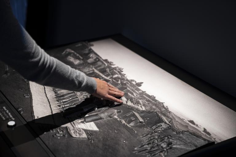 A hand touching a large image that is raised in places, and the people and buildings portrayed all have visible texture.