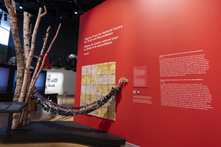 A museum exhibit featuring many traditional Indigenous medicine bags hanging from a baby swing. One end of the swing is attached to birch tree trunks and the other to a red wall that includes text panels. Partially obscured.