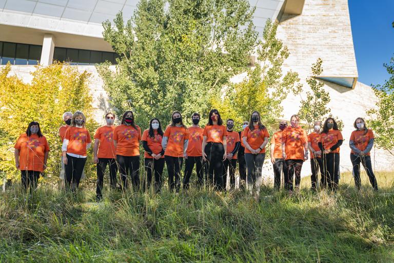 People wearing orange T-shirts stand in front of a row of trees. There is long grass in the foreground. The T-shirts bear images of small hand prints grouped in a heart shape with the number 215+ above them. Partially obscured.