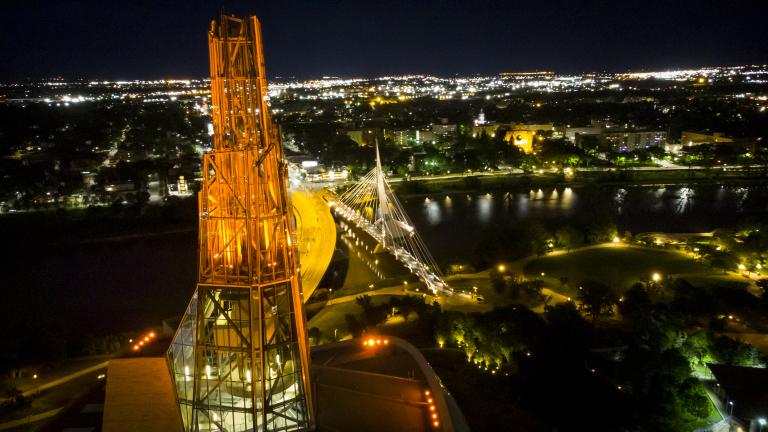 A glowing orange tower is pictured from above against a background night-time cityscape with view of a river and a bridge.