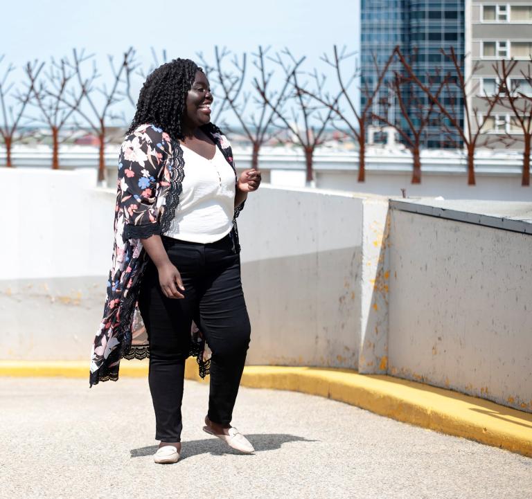 A smiling Black woman stands in front of a row of trees. She is looking to her left. Partially obscured.