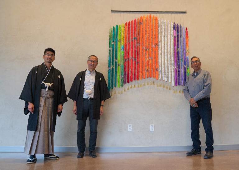 Three men stand beside a colourful artwork made of one thousand folded paper cranes. Two of the men are wearing traditional Japanese clothing. The other man is wearing a jean shirt and bolo tie.