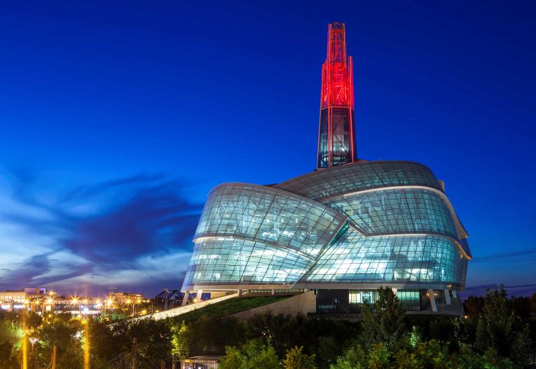 A huge building with a rounded glass façade is topped by a glowing red tower. Partially obscured.