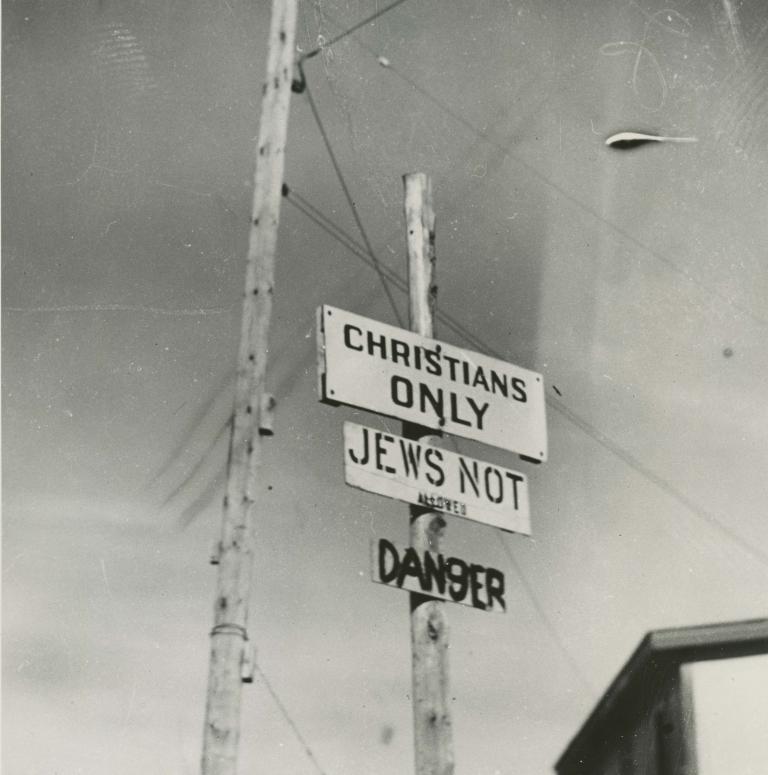 Photo noir et blanc de trois écriteaux fixés sur un poteau, où l’on peut lire en anglais « Chrétiens seulement », « Interdit aux Juifs » et « Danger ». Partially obscured.