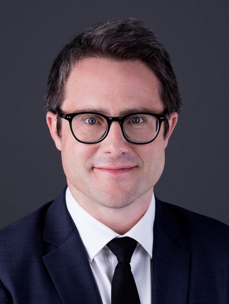 Headshot of a smiling man with short, dark-brown hair. He is wearing glasses and a suit.