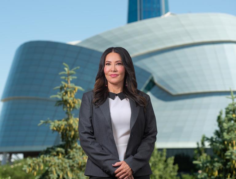A woman with long dark hair, wearing a suit jacket, stands with her hands clasped in front of her. Behind her is a large, unusual building with a rounded glass façade and a tower. Partially obscured.