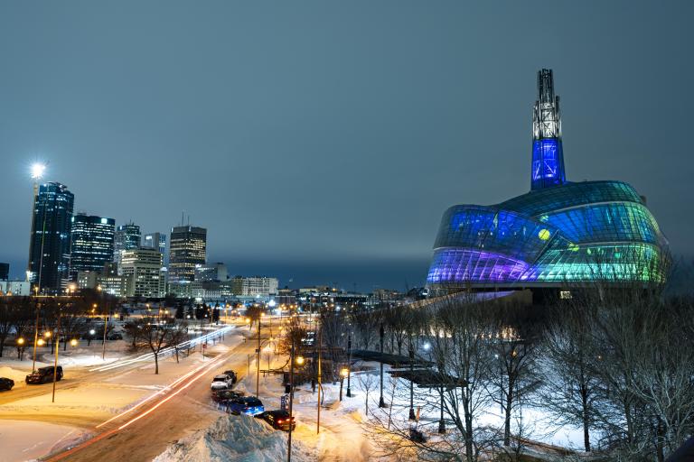 The CMHR’s exterior is seen at night with its glass panels lit up blue, green and purple. Partially obscured.