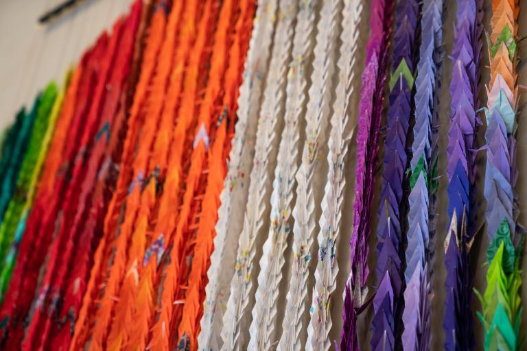 Multi-coloured origami cranes strung together vertically and hung on a wall. Partially obscured.