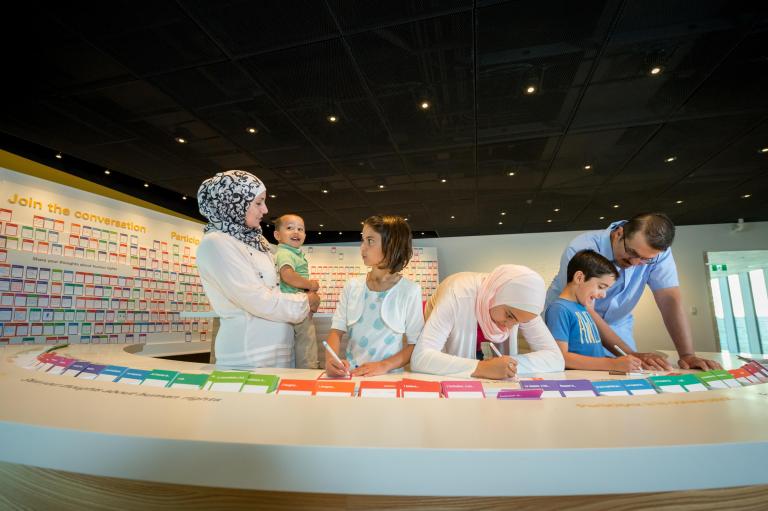 A family of six stands in front of curved table with coloured cards on it. Three family members are writing on cards with markers while the woman holds a smiling toddler. Partially obscured.