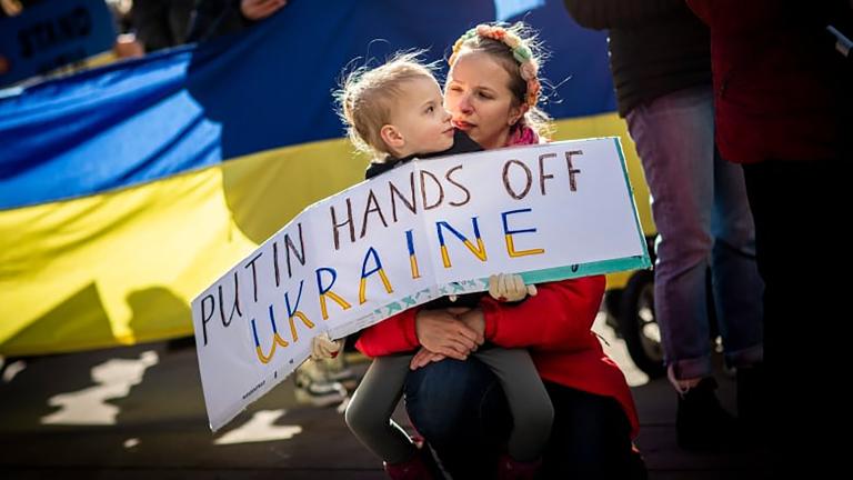 A child is holding a protest sign that says: “Putin, hands off Ukraine.” The child is sitting on their caregiver’s lap. Behind them is a Ukrainian flag. Partially obscured.