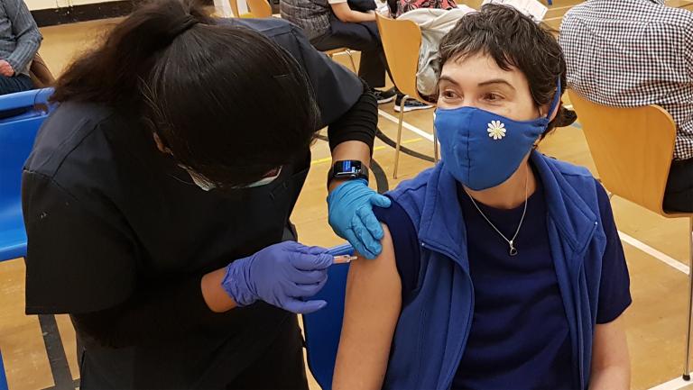 A healthcare worker prepares to give a vaccine to a patient. Both people are wearing medical face masks. Partially obscured.