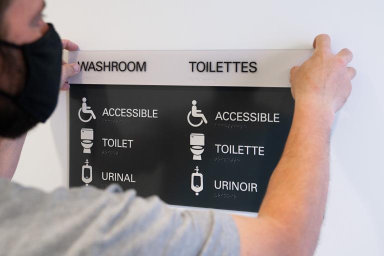 A man holds up a sign that says "Washroom" as it is mounted to a wall. Underneath the title, the words "accessible," "toilet" and "urinal" are accompanied by descriptive icons. Partially obscured.