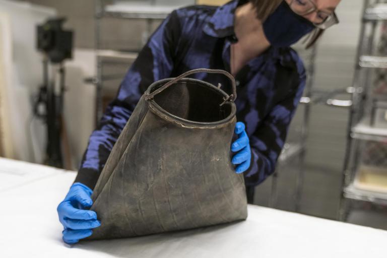 A woman holds a scuffed black rubber bag with a roughly shaped curved metal handle spanning the top.