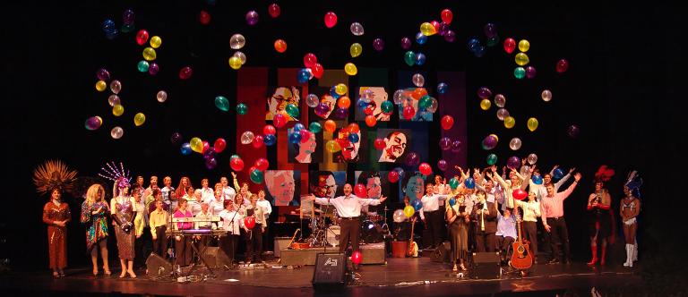 Rainbow Harmony Project Choir singing on a wooden stage inside a church. Partially obscured.