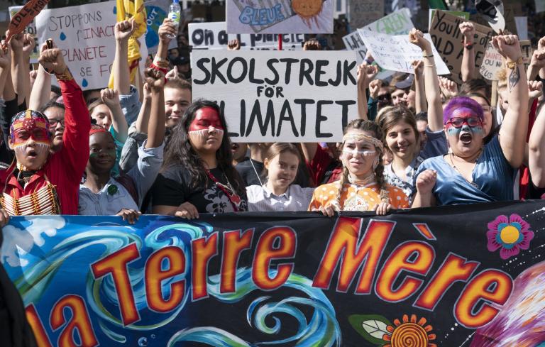 A crowd of youth hold protest signs and stand behind a large banner that reads “La Terre Mère,” or “mother earth” in English. Partially obscured.