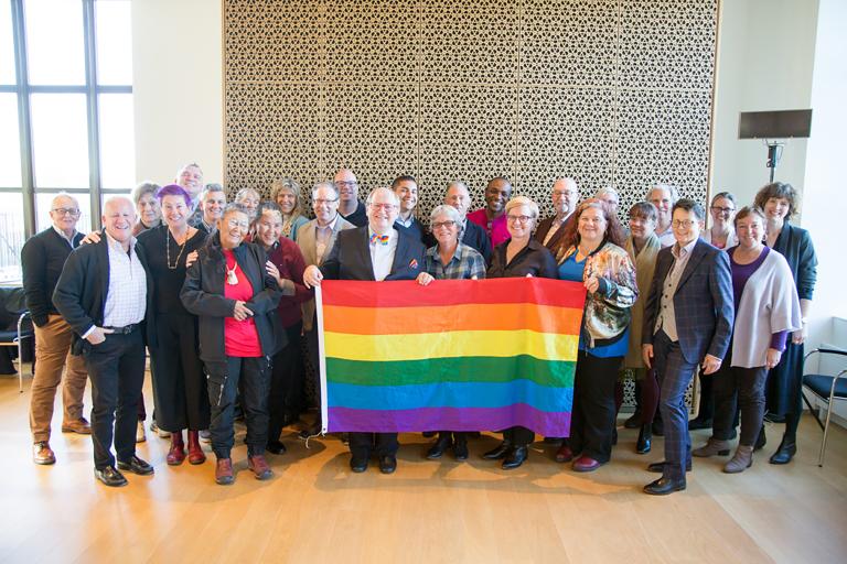 Twenty-eight members of the LGBT Purge Fund Board and LGBTQ2+ National Monument Advisory Committee standing together and holding a rainbow flag, at the first Monument visioning session in 2019. Partially obscured.