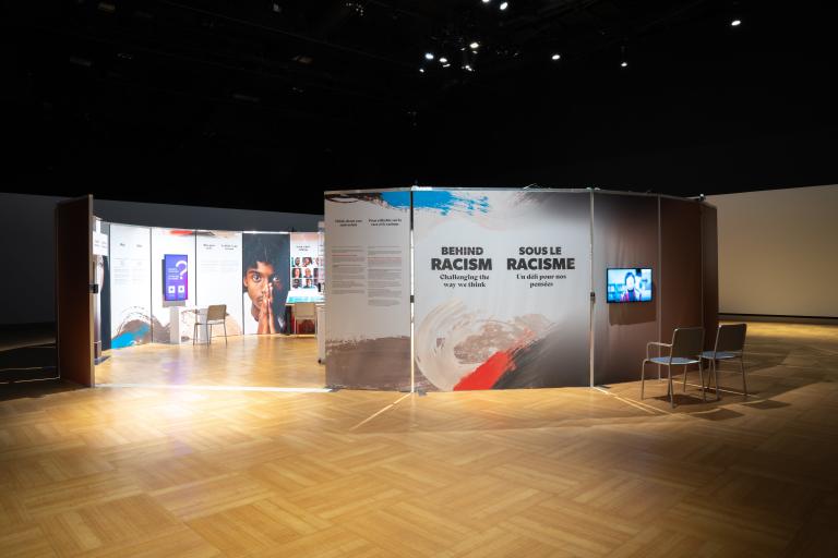Upright panels are arranged in a circle in a dimly lit room. A gap offers people a way to enter the circle, and the interior of the circle is lit brightly. Next to the opening, a panel shows the words "Behind Racism: Challenging the Way We Think." Partially obscured.