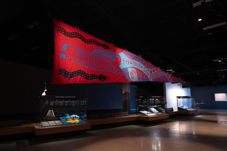 Une grande bannière rouge est suspendue dans la galerie au-dessus de trois vitrines. En français, les mots en squamish sur la bannière signifient : « Protéger cette terre, c’est dans notre sang ». Partially obscured.