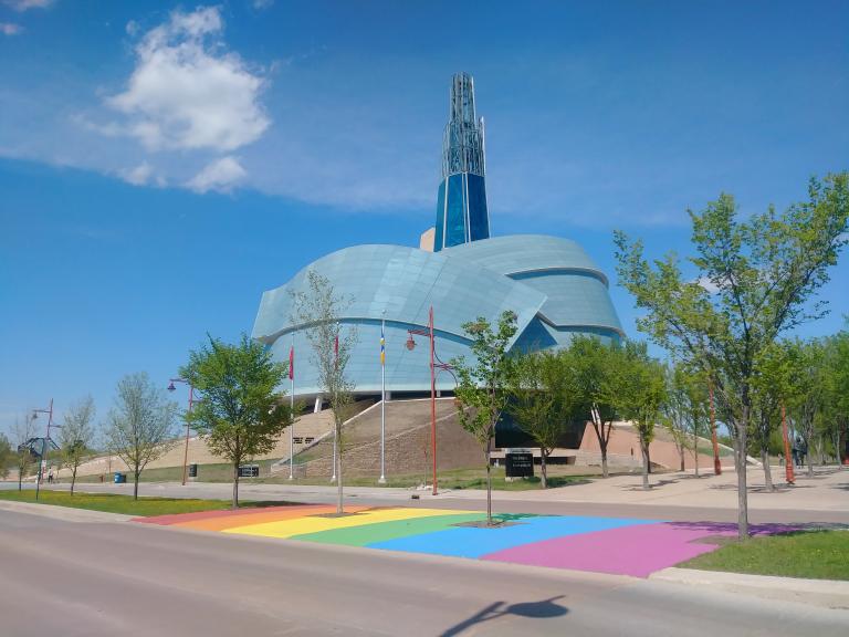 A rainbow crosswalk in front of the Canadian Museum for Human Rights on a sunny day Partially obscured.