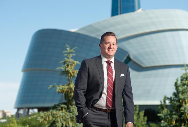 A person standing with the Museum in the background. He is wearing a suit, smiling, with his right hand in his pocket, looking directly at the viewer. Partially obscured.