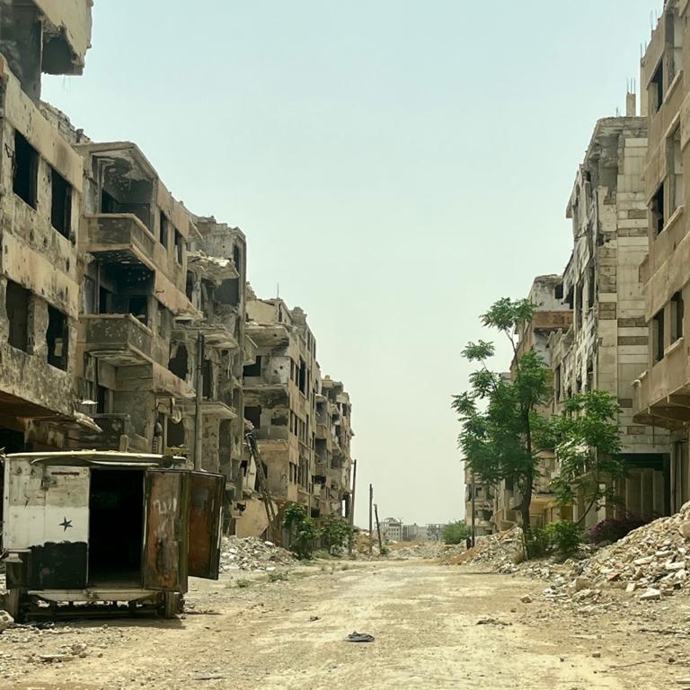 An abandoned city street where the building windows are gone, rubble lies piled in the road and a truck sits abandoned with its back doors open. Partially obscured.