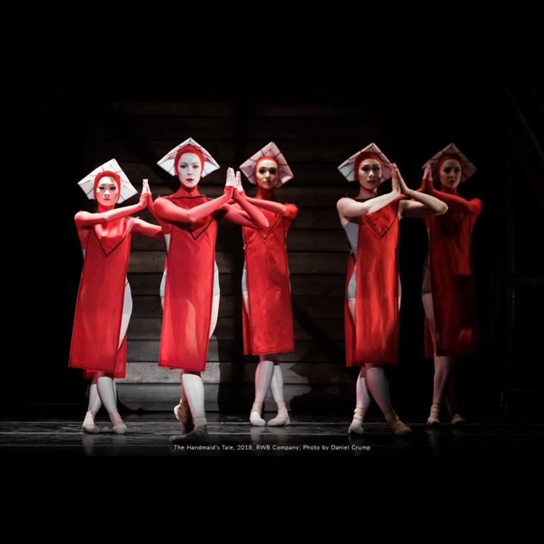 On a dark stage are five ballet dancers dressed in red knee-length flowy costumes with white square-like hats. They are looking forward with their hands in prayer. Partially obscured.