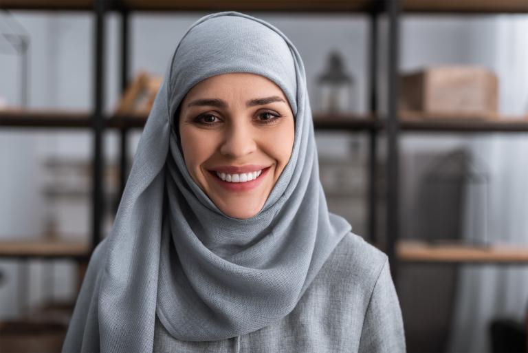 A woman wearing a blue hijab smiles toward the camera, with a bookshelf in the background. Partially obscured.