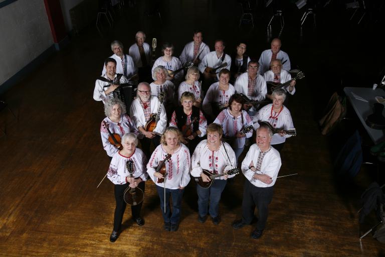 A group of twenty-two people facing the camera and smiling. They are all wearing dark-coloured pants and white shirts with red embroidery. A majority of the group are holding their instruments, mainly mandolins. Partially obscured.