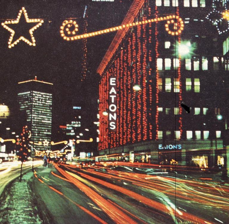 Late 1960s-era photo of downtown Winnipeg showing the Eaton’s building decorated with many bright Christmas lights. Partially obscured.