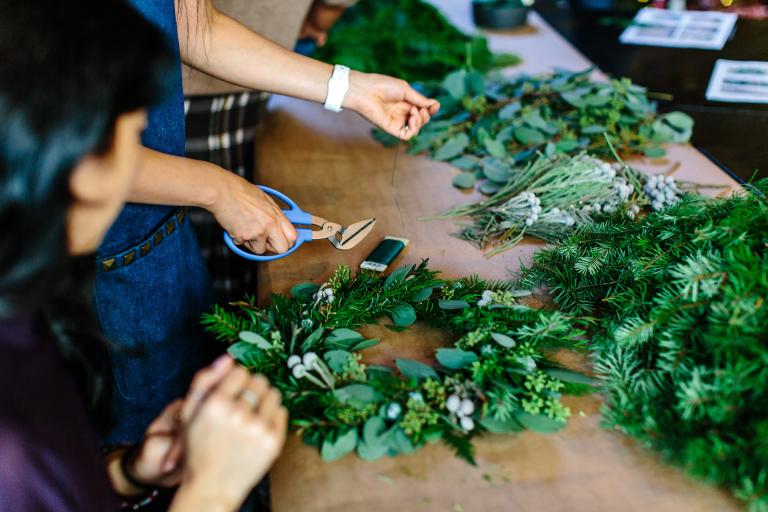 A close-up of two people making a wreath. One person is holding clippers and is about to cut some wire. Partially obscured.