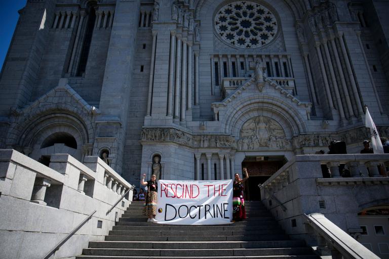 Two people in braids and ribbon skirts raise fists and hold a large cloth banner reading “RESCIND THE DOCTRINE” on the steps of an enormous cathedral. Partially obscured.
