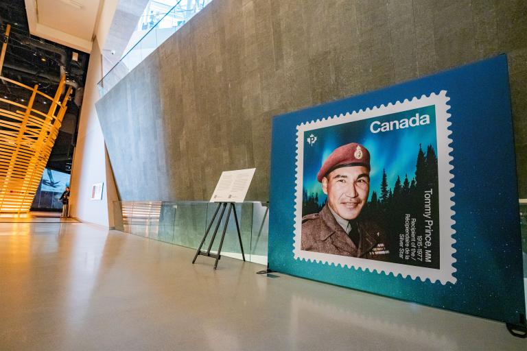 An enlarged Canadian stamp showcases a man with a red military beret with an aurora borealis and darkened forest in the background. Partially obscured.