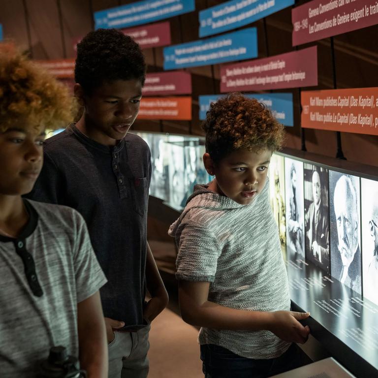 Three young children look at photographs and a text display. Partially obscured.