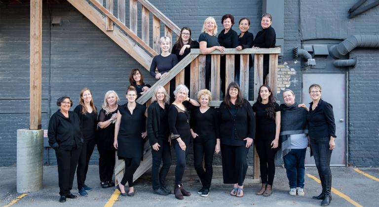 Women’s choir members are dressed in black and posed along an outdoor wooden staircase. Partially obscured.