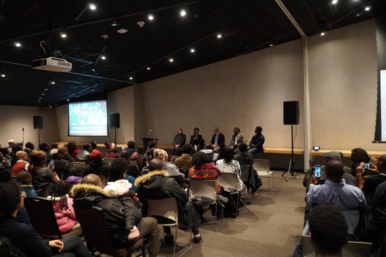 A panel of five people are seated in front of a large crowd inside the Canadian Museum for Human Rights. Partially obscured.