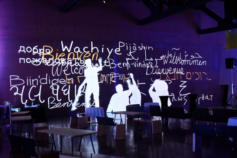 Interactive screen depicts human figures writing “welcome” in several different languages. Partially obscured.