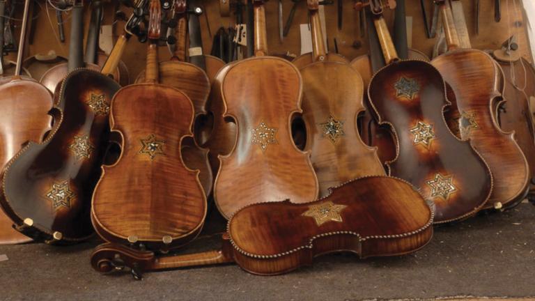 Several restored violins are pictured leaning on each other in a pile. Each of the violins carry at least one Star of David Partially obscured.