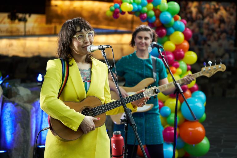 Two guitar players in bright and colourful outfits sing into microphones in front of a balloon arch. Partially obscured.