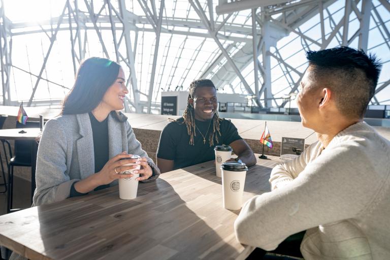 Three people sit around a table, smiling, each with a paper coffee cup in front of them. The sun shines behind them, through the panes of glass that wrap around the Canadian Museum for Human Rights. Partially obscured.