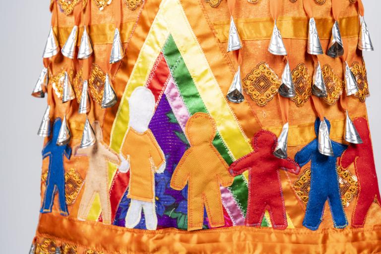 A section of an orange jingle dress is shown. Jingles, or small silver cone-shaped pendants, are visible at the top of the image. Under them, seven human figures are shown, all in various colours and sizes. One of the figures is wearing an orange shirt. These human figures are holding hands inside a multicoloured tipi. Partially obscured.