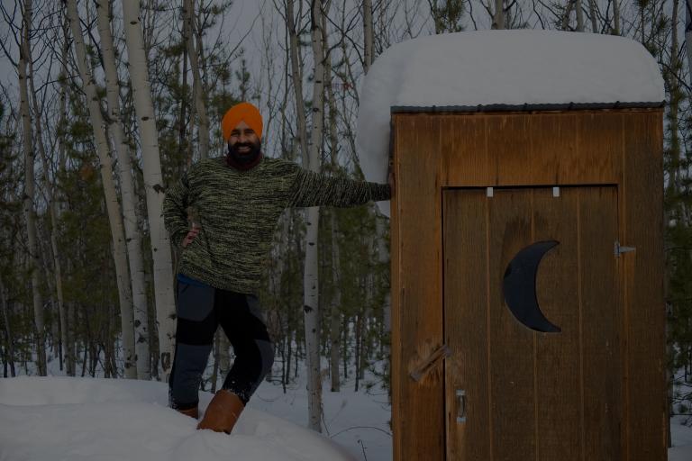 A Sikh-Canadian man, smiling, wearing a green sweater, blue and brown pants, boots, and an orange turban, leaning against a cabin with a crescent moon. Partially obscured.