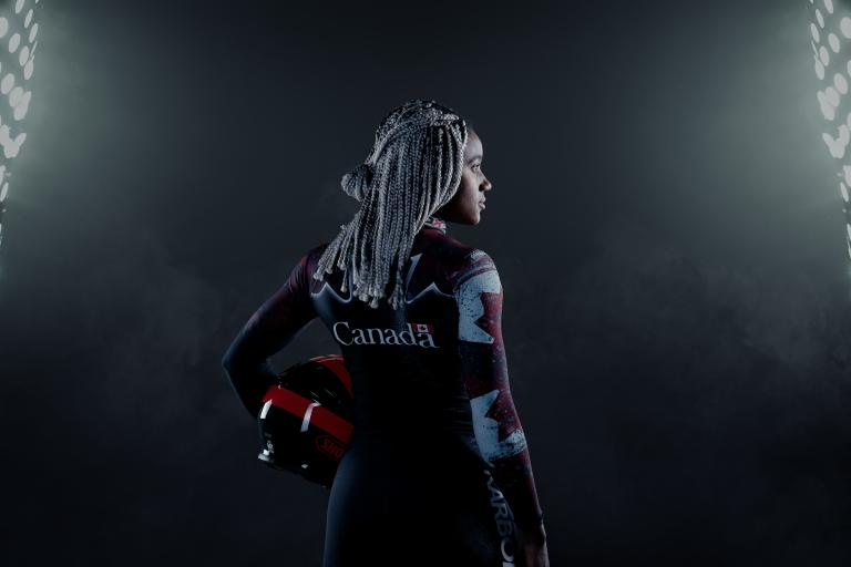A black woman stands in a dark place between two large projectors. She's holding a helmet and wears a Canadian bobsleigh suit. Partially obscured.