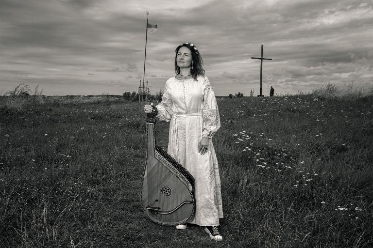 A woman in a long white dress stands in a field holding a bandura, a Ukrainian plucked string folk instrument. Partially obscured.