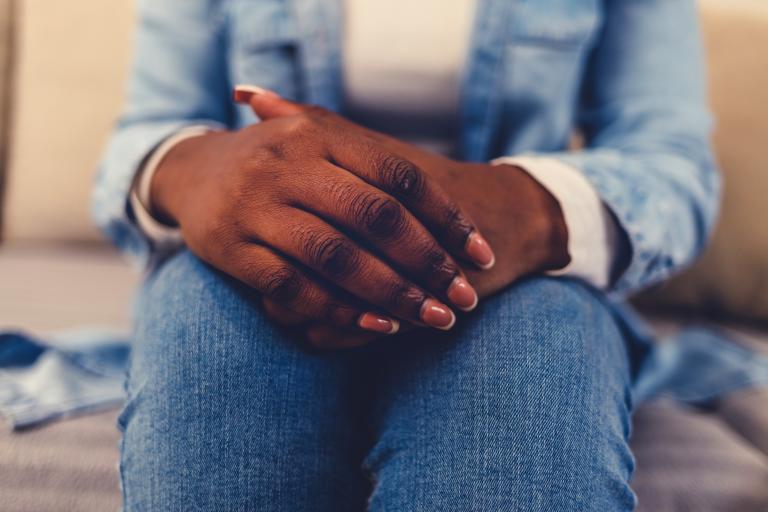 Close-up of a dark-skinned person sitting on a couch, with their hands clasped on their lap. Partially obscured.