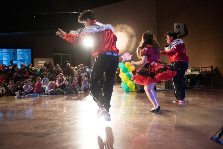 Three dancers in red and white outfits perform in front of a crowd of children at the Canadian Museum for Human Rights. Partially obscured.