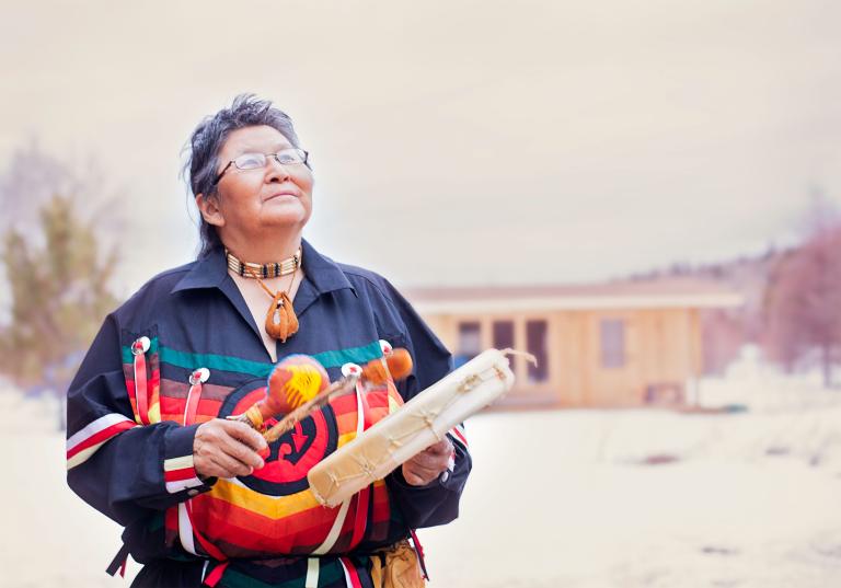 Elder Ma-Nee Chacaby stands outside surrounded by trees and cabin looking up at the sky playing a drum. Partially obscured.