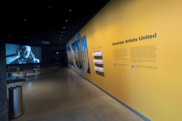 A yellow wall with the words "Ukrainian Artists United" and a dynamic blue and yellow graphic is right side of the image. On the left side, an image is projected on a screen showing a person with headphones and an open mouth, as though they are singing. Benches for sitting are placed in front of the screen. Partially obscured.