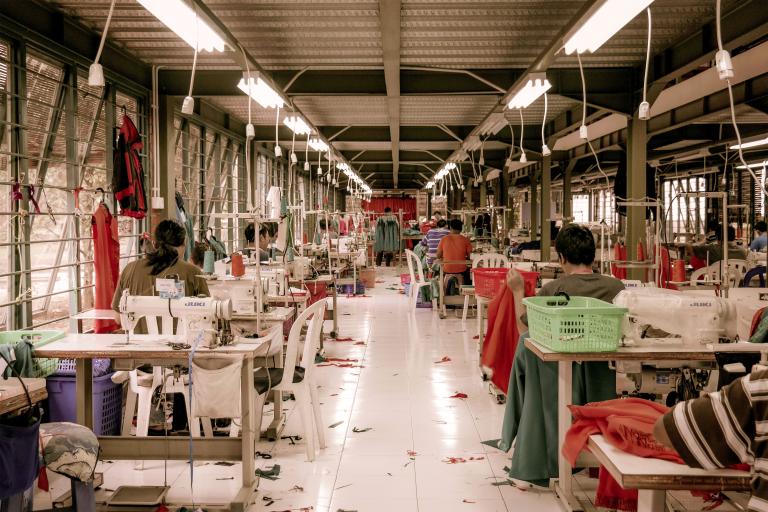 Garment workers sit at desks with sewing machines in a warehouse. Partially obscured.