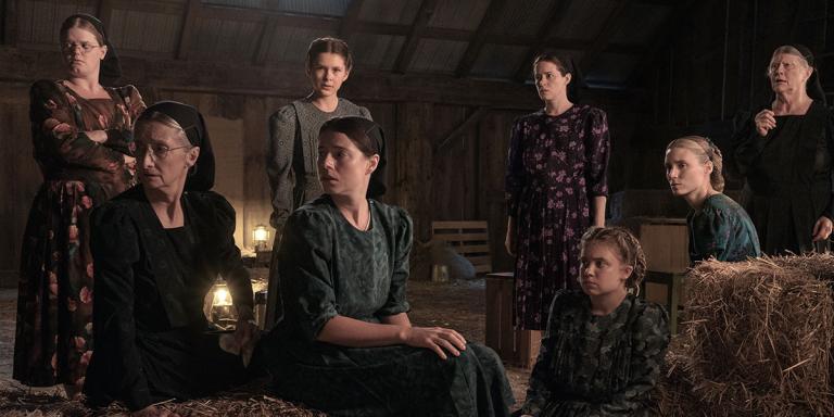 A group of women dressed in head coverings and long dresses are gathered in a barn filled with hay bales and lit by lanterns. Some of them standing and some are sitting while they look to the left. Partially obscured.