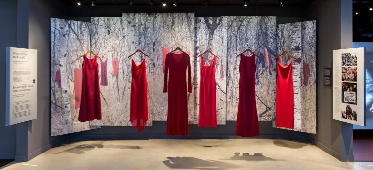 Five red dresses, representing missing and murdered Indigenous women and girls, hang in a row in the Canadian Journeys gallery as part of an exhibit titled “From Sorrow to Strength.” Behind the dresses, the scene of a snow-covered forest with more red dresses hung on the branches of trees is displayed on a series of banners. Partially obscured.